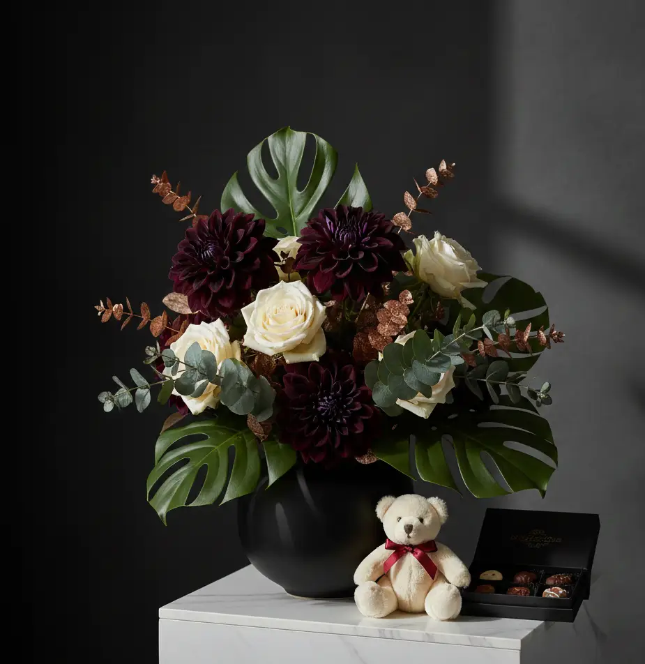 Luxury gift set by Konceptify featuring a bespoke floral arrangement of midnight plum dahlias, cream roses, and monstera leaves, paired with a plush teddy bear and gourmet chocolates on a marble pedestal. Bespoke Floral Designer in Durban